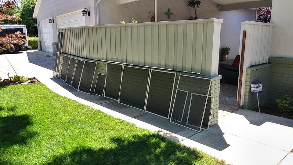 Several dark framed window screens leaning against a light green metal fence in front of a house.