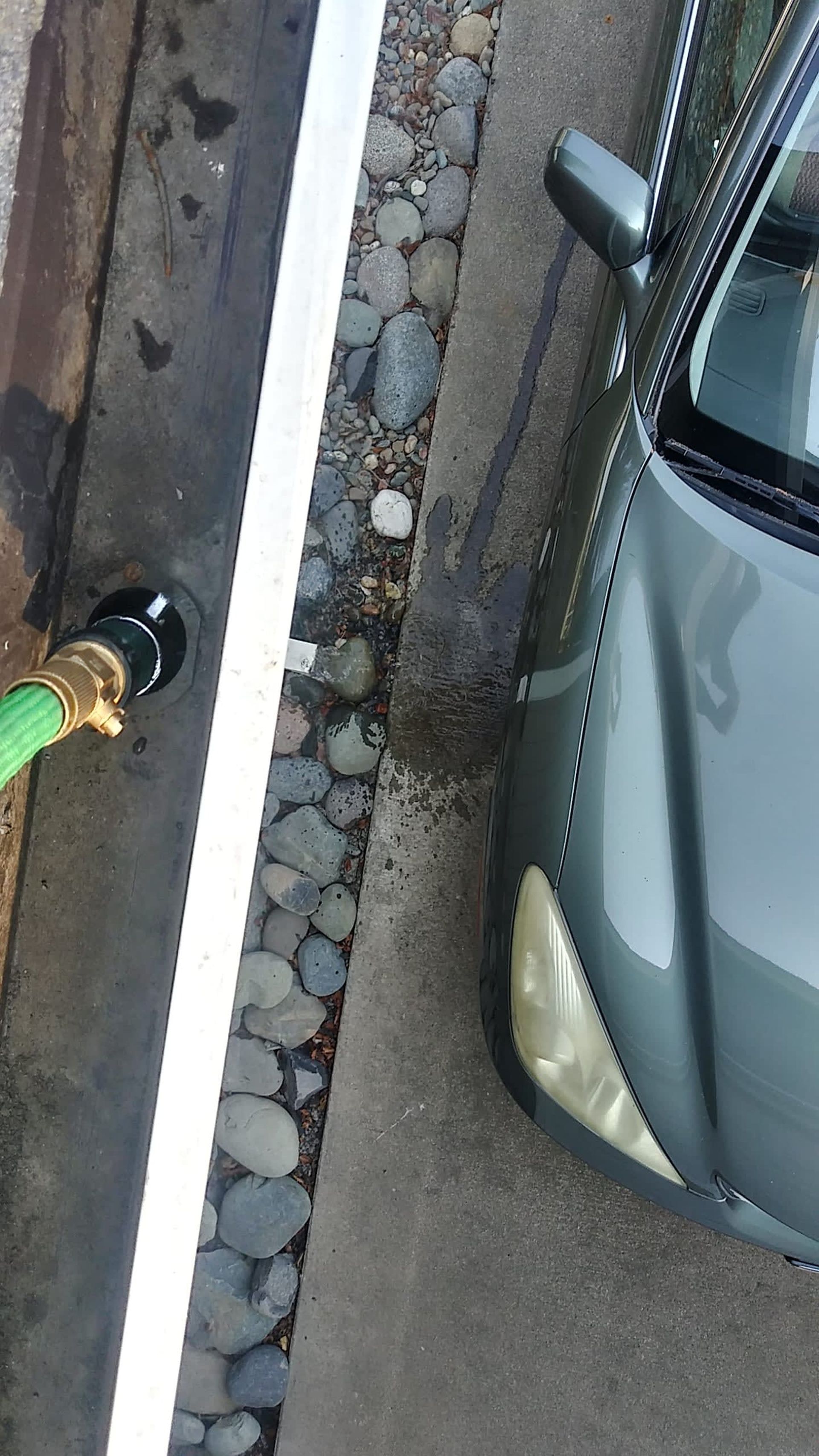 A green car next to a curb with a white line and gravel, being washed with a hose.