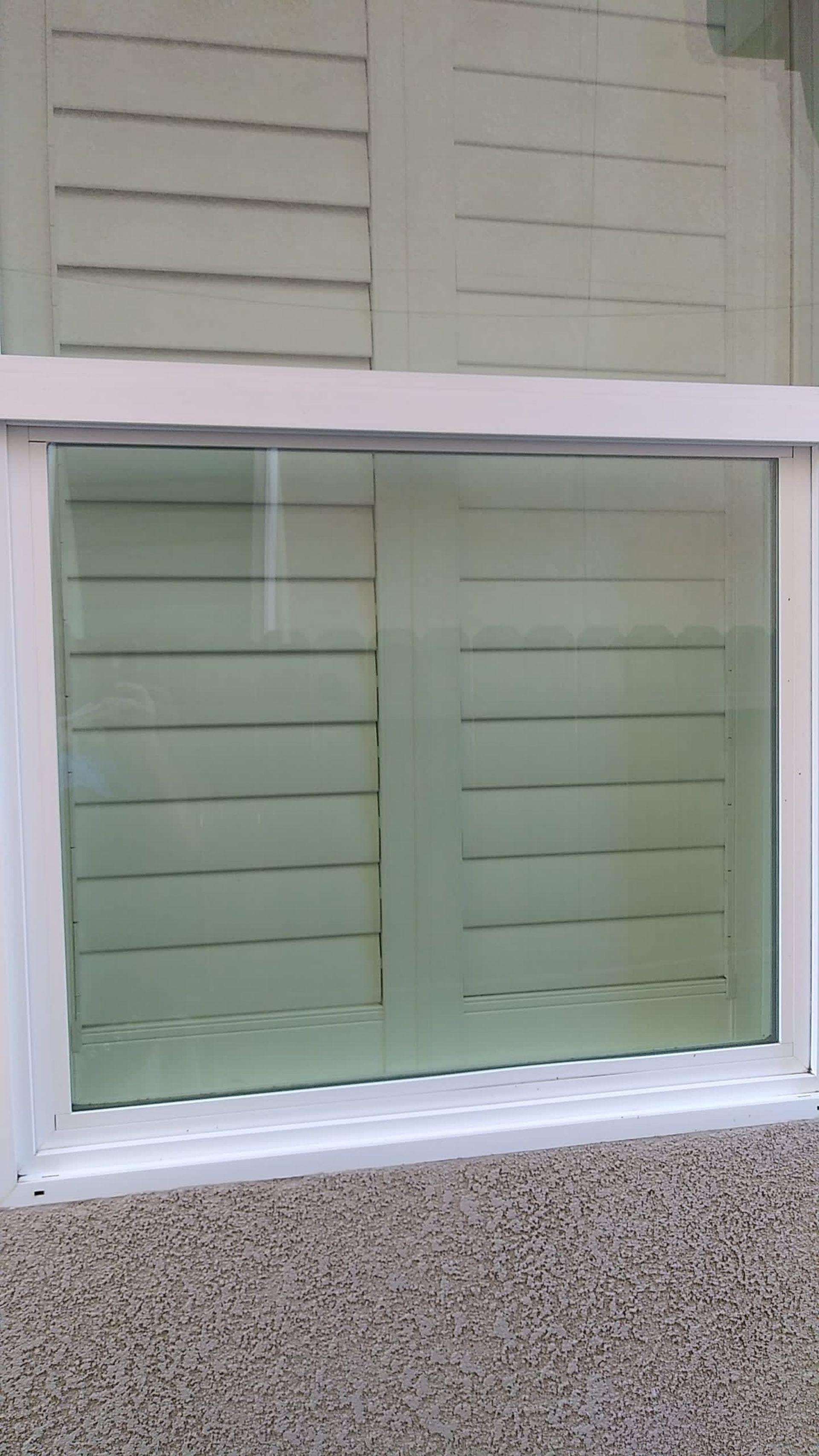 White-framed window with view of light green, slatted shutters. Mounted on a gray textured exterior wall.