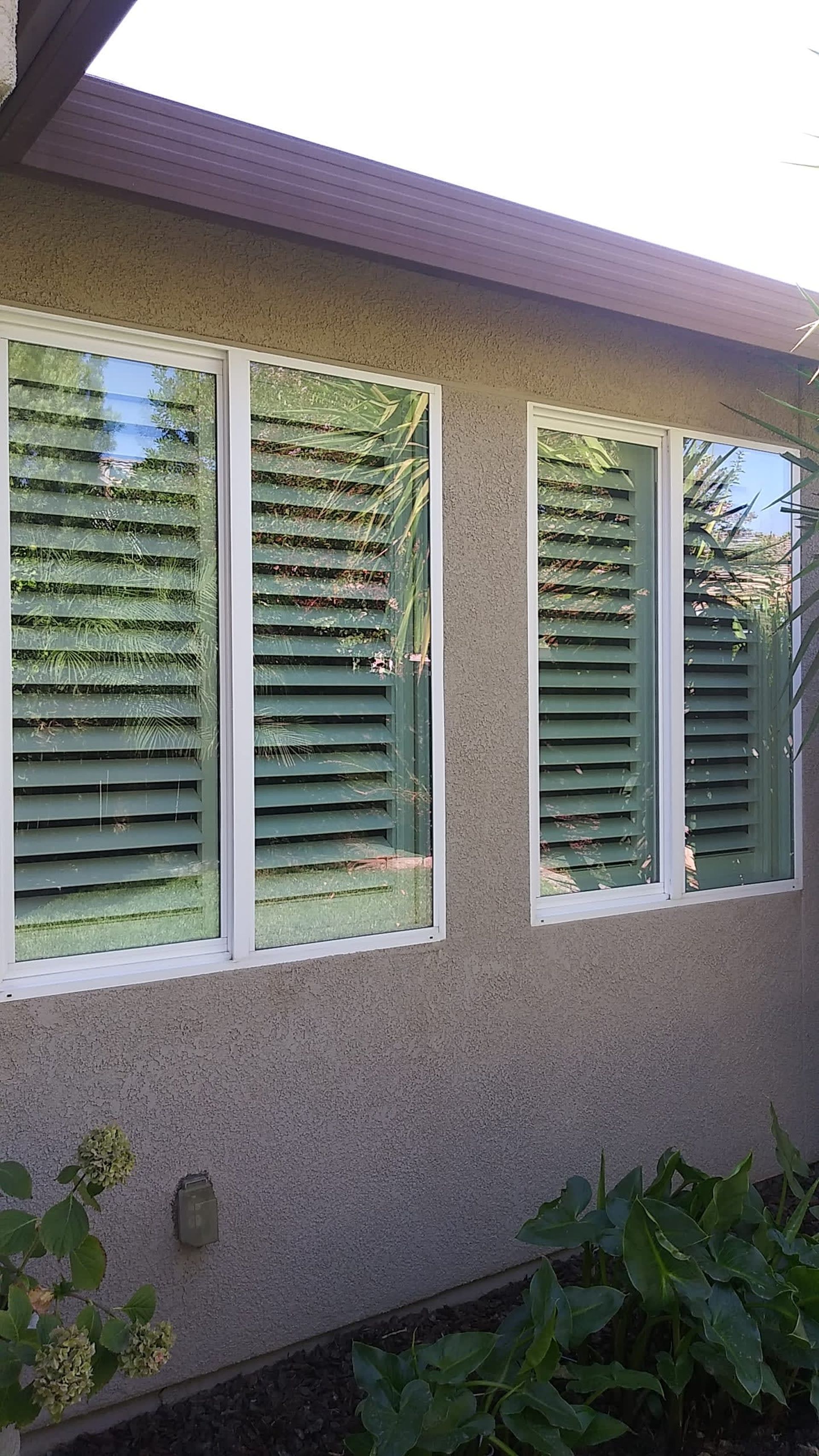 Exterior view of three windows with closed, horizontal-slat shutters, set in white frames, against a stucco wall.