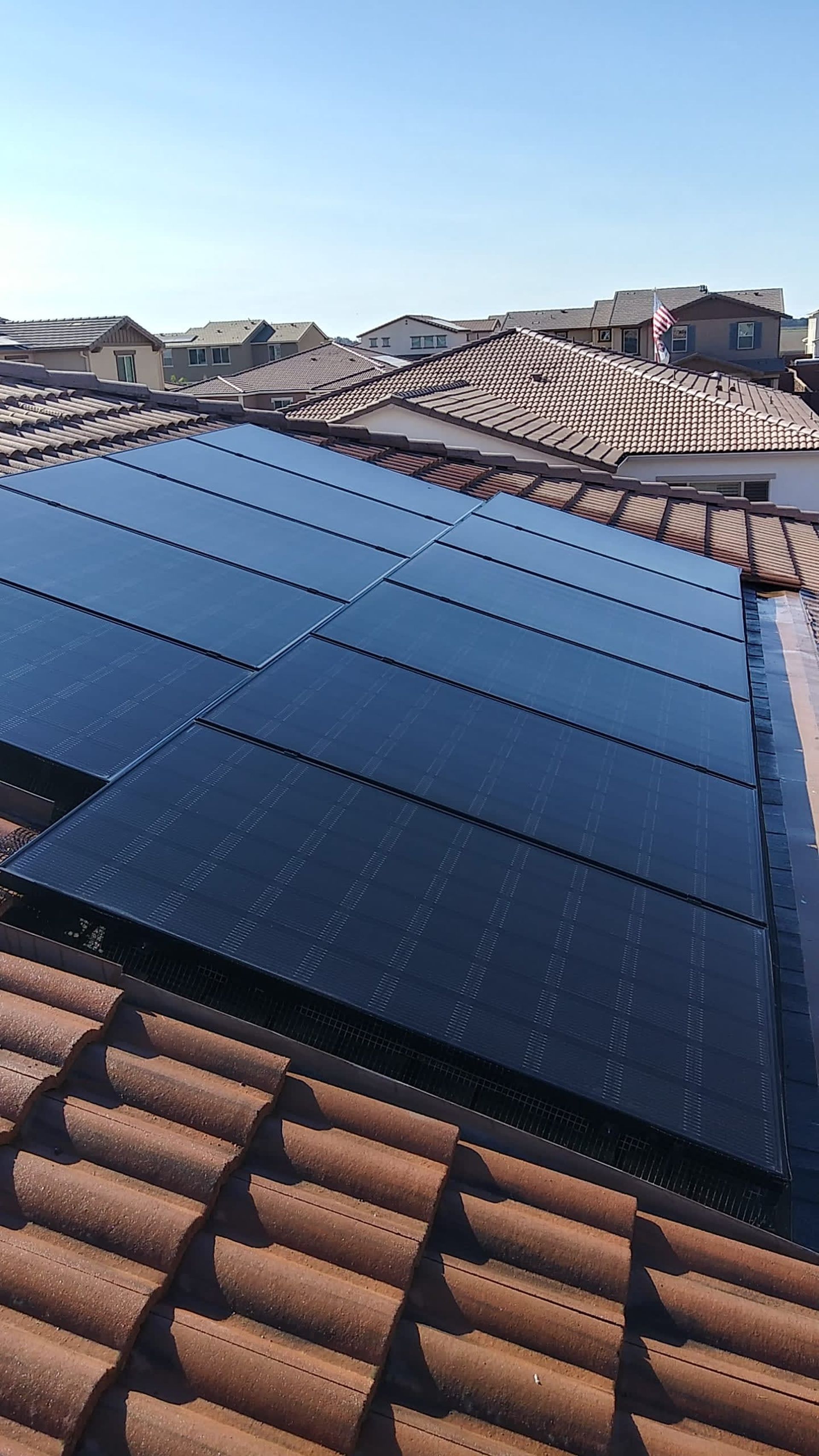 Solar panels on a terracotta tile roof against a backdrop of similar rooftops under a clear blue sky.