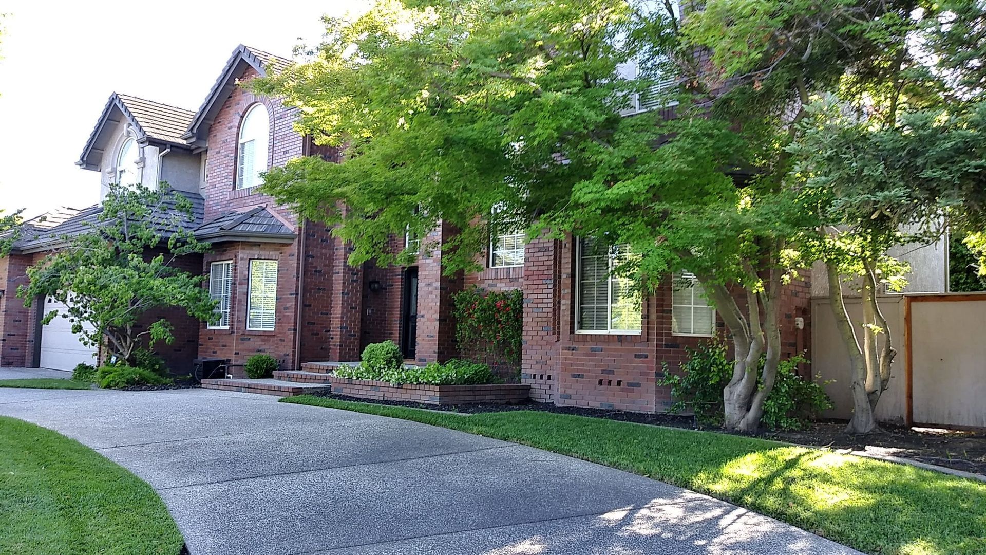 Brick house with a long, gray driveway and lush green trees in the front yard.