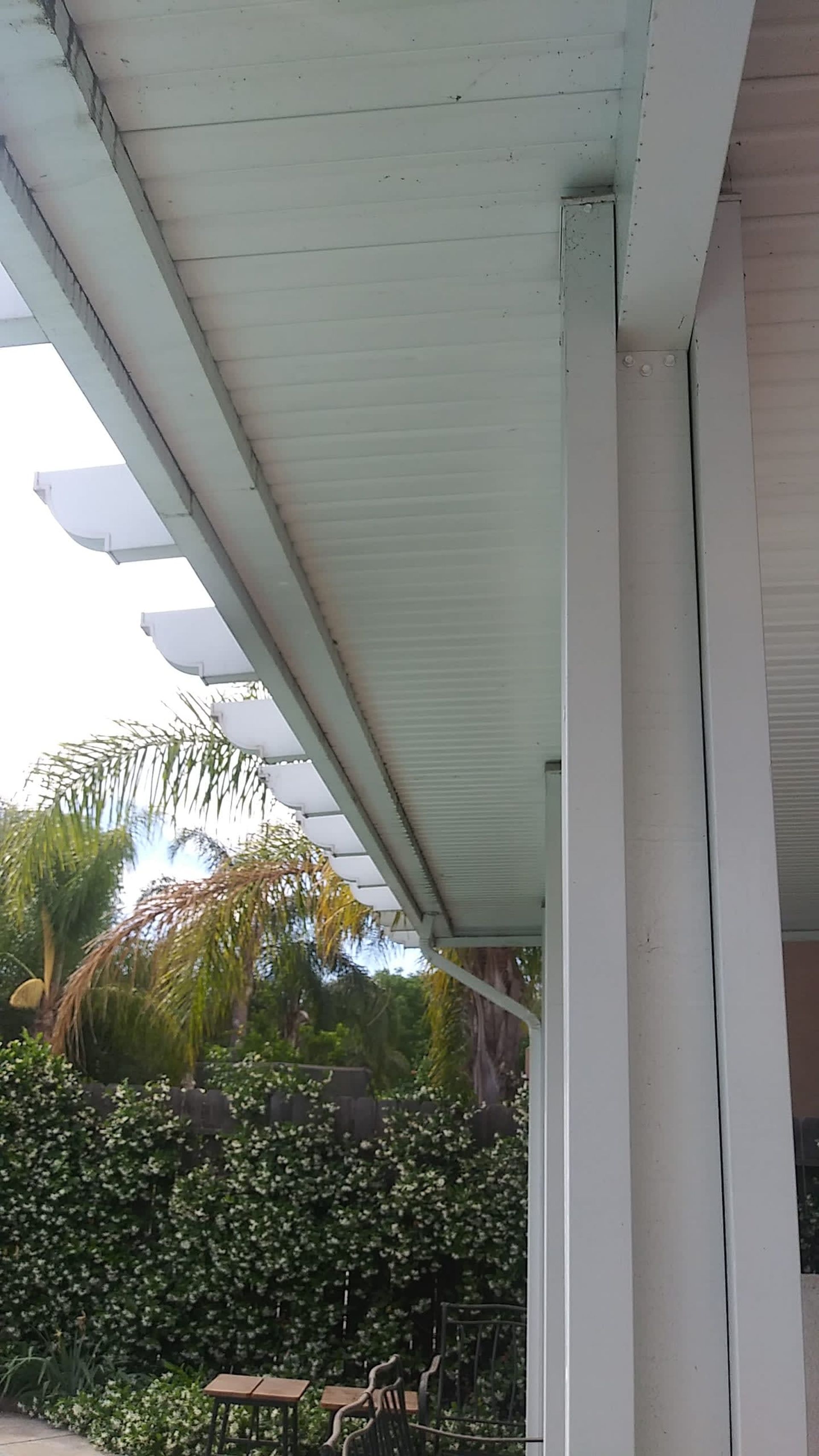 White pergola with latticework, side view. Overhanging roof supported by a column next to a wall, greenery in the background.