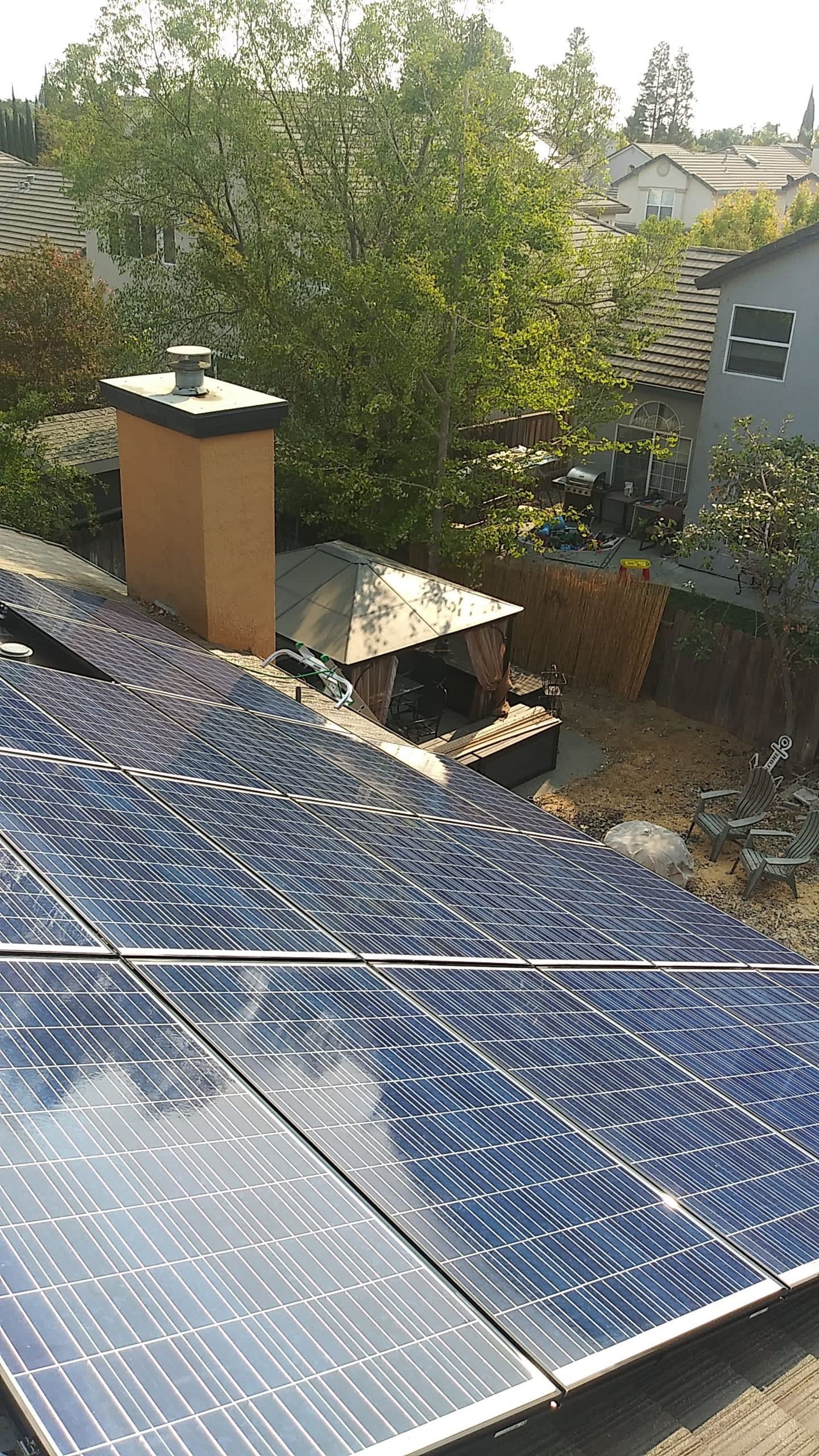 Solar panels on a rooftop with a chimney, trees, and other houses in the background.