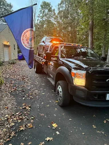 Black utility truck with flashing lights carrying a blue flag beside a house on a leaf-covered driveway