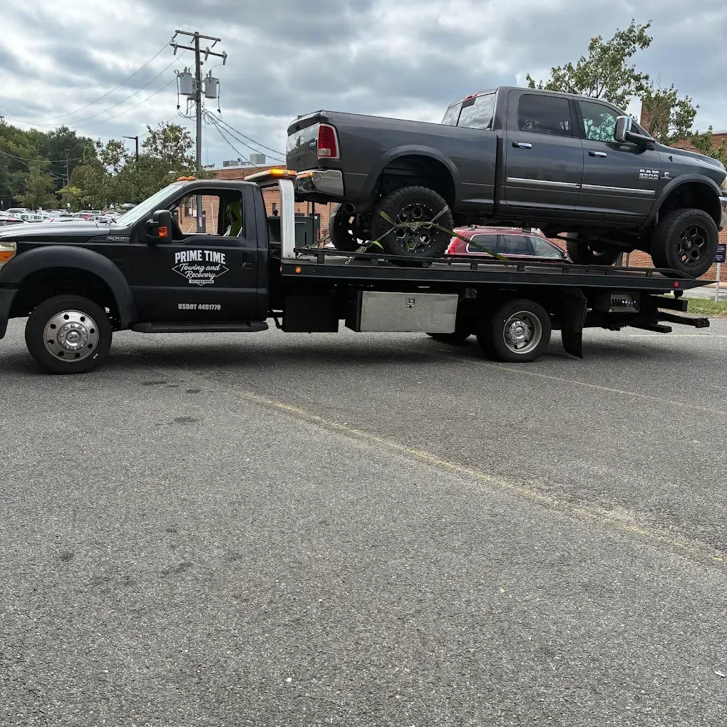 Tow truck carrying a lifted dark pickup on a flatbed in a parking lot under cloudy skies