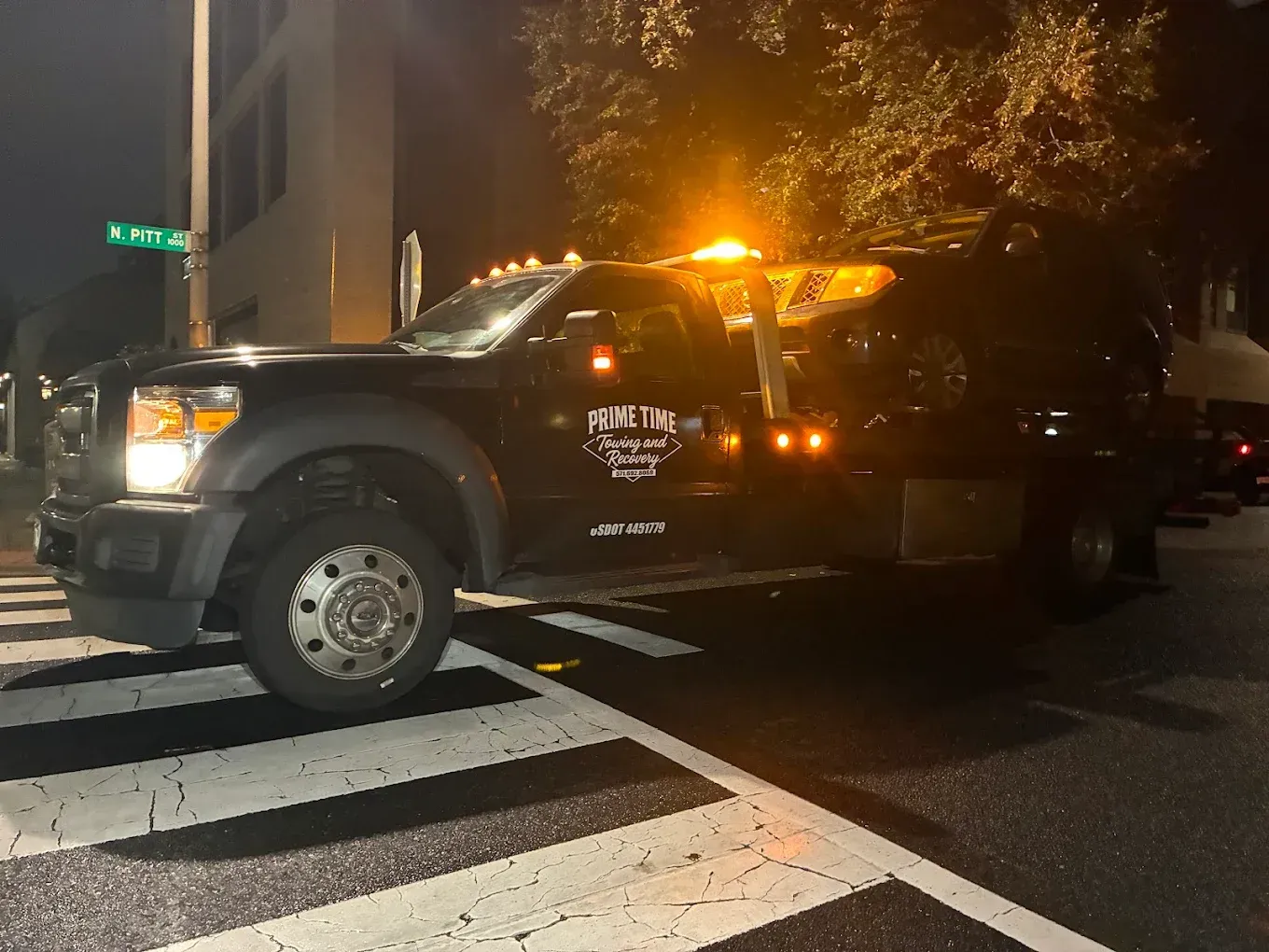Parked black tow truck with flashing amber lights at a crosswalk at night
