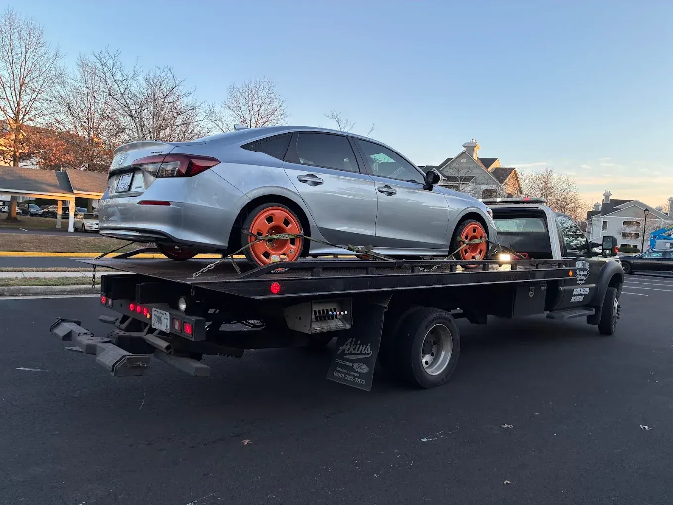 Silver sedan with orange wheels on a flatbed trailer in a suburban street at dusk
