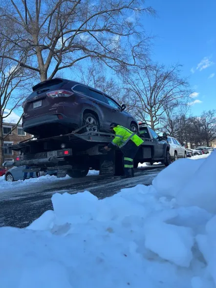 Tow truck lifting a dark SUV in a snowy street as a worker in a neon jacket stands beside it.