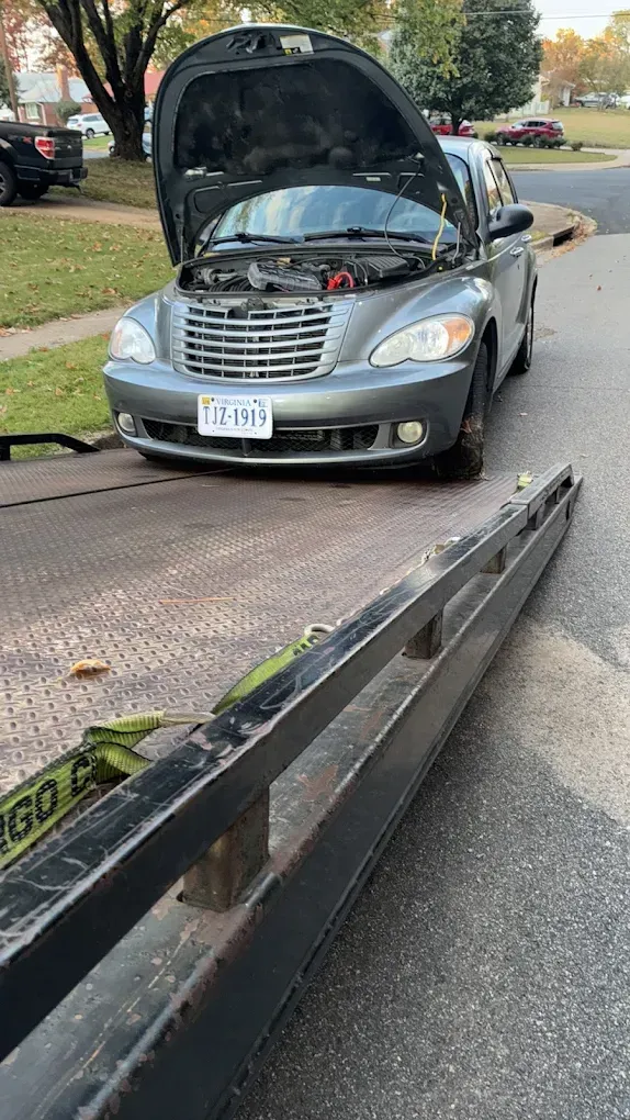 Silver car with raised hood after roadside breakdown, near a tow truck ramp on a suburban street