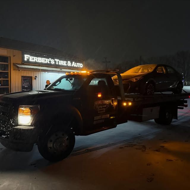 Tow truck hauling a black car at night outside Ferber’s Tire & Auto.