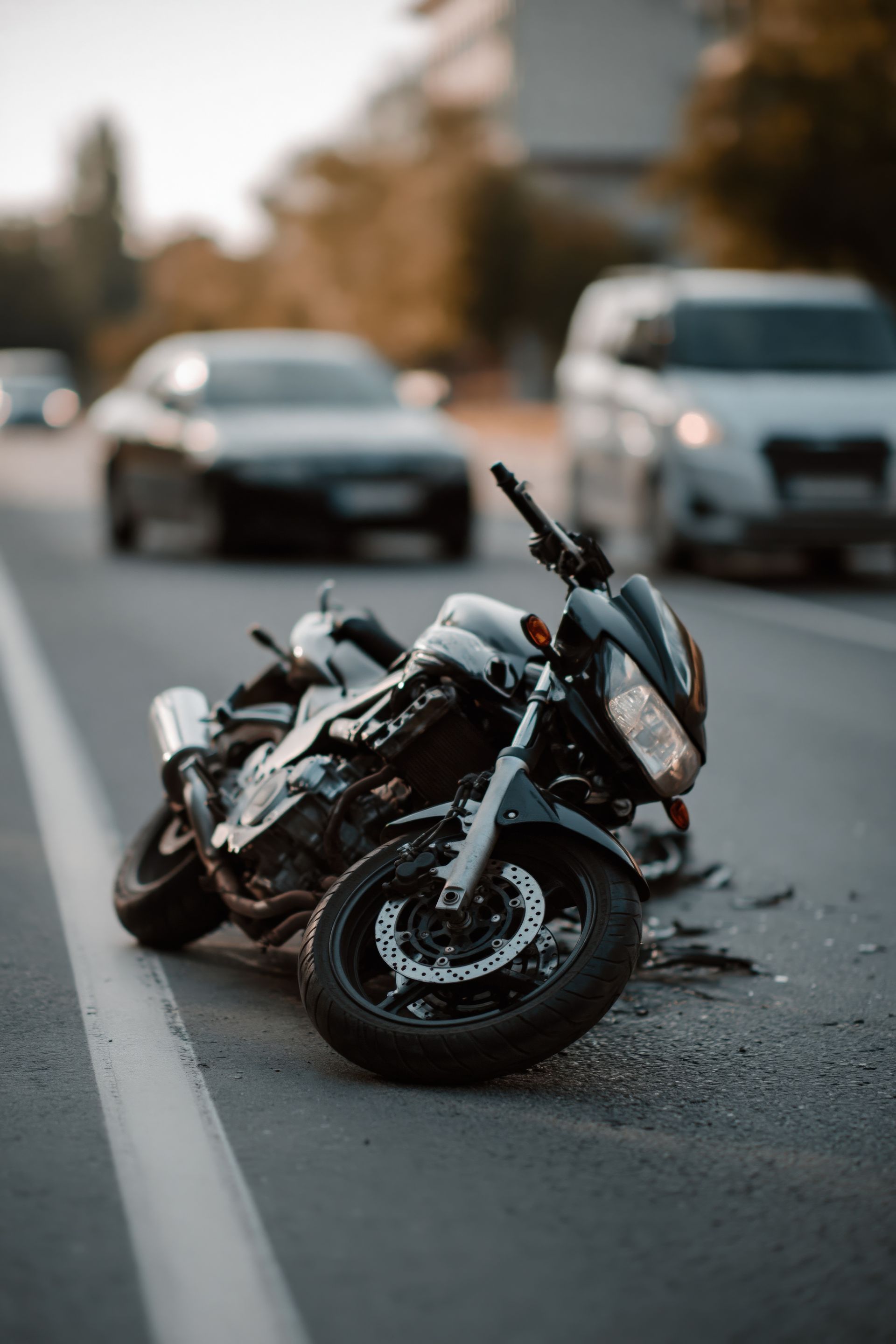 Motorcycle lying on its side in a road lane, with cars approaching in the background.