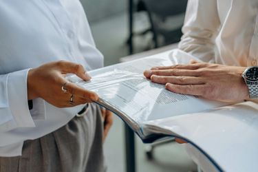 Two people reviewing documents, with hands visible on a binder, wearing white shirts and gray pants.