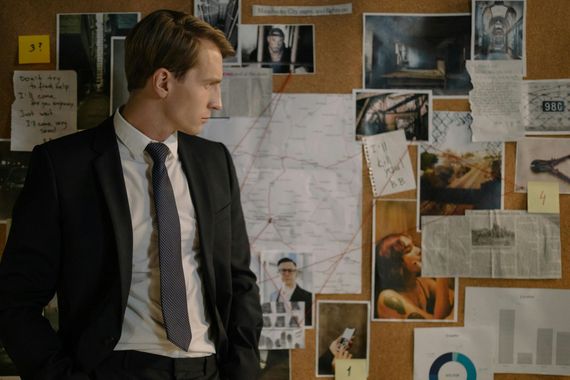 A man in a suit stands looking at a corkboard covered in evidence, maps, and photos. He appears focused.