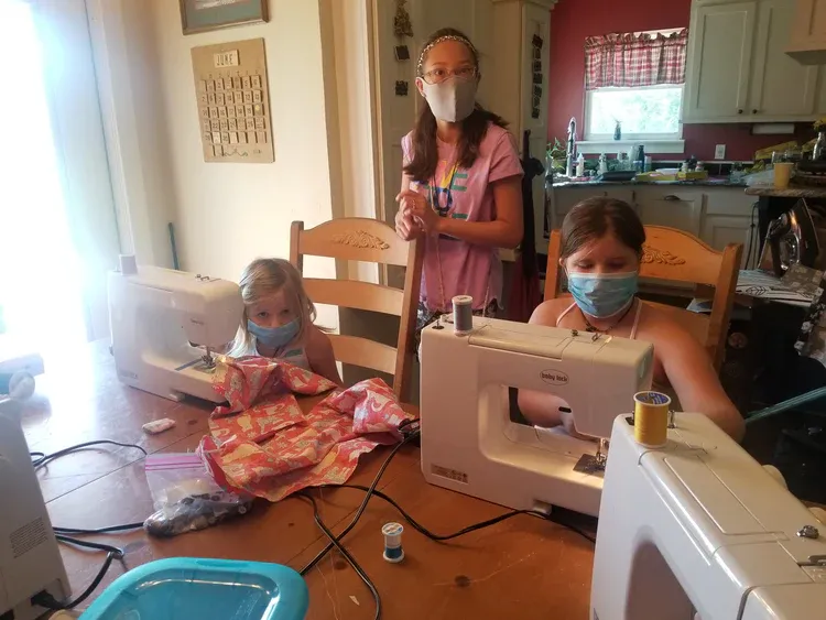 Two girls wearing face masks are sitting at a table with sewing machines.