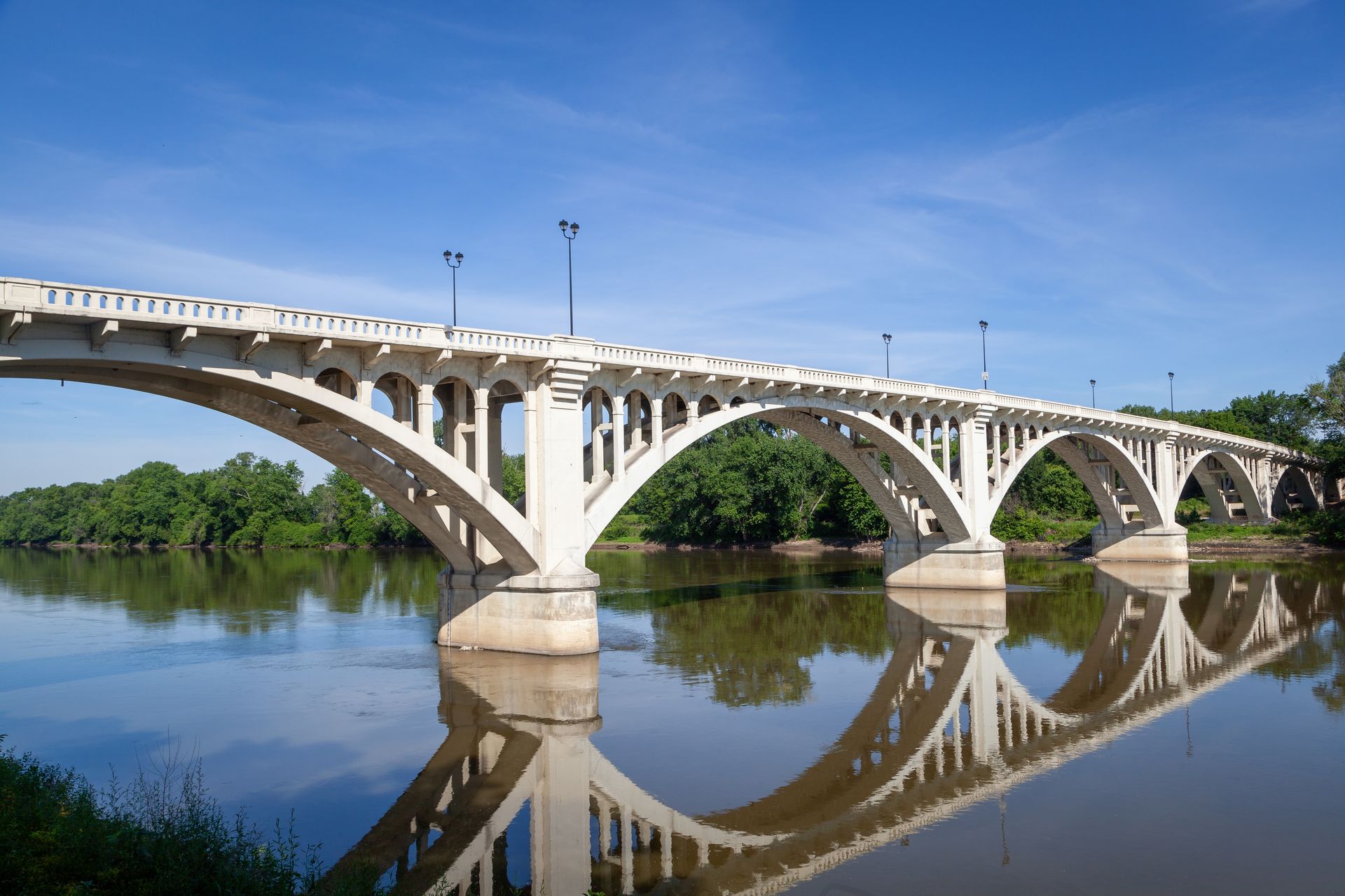 White arched bridge over calm river, reflecting in the water, blue sky.