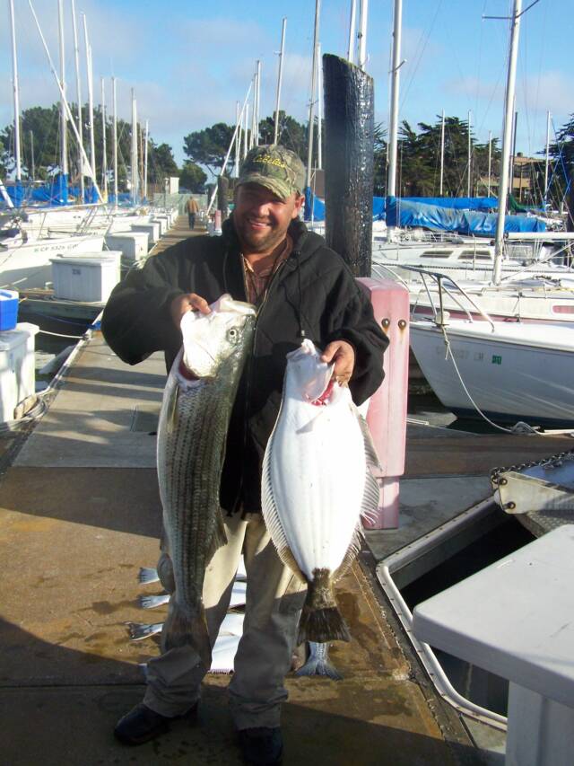 A man is holding two large fish in front of boats