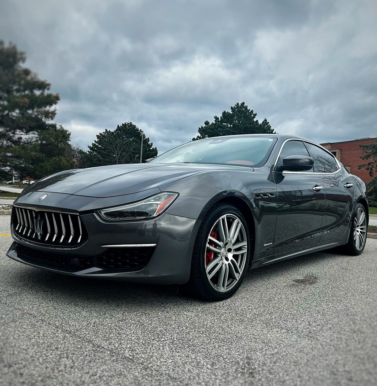 A gray maserati ghibli is parked in a parking lot on a cloudy day.