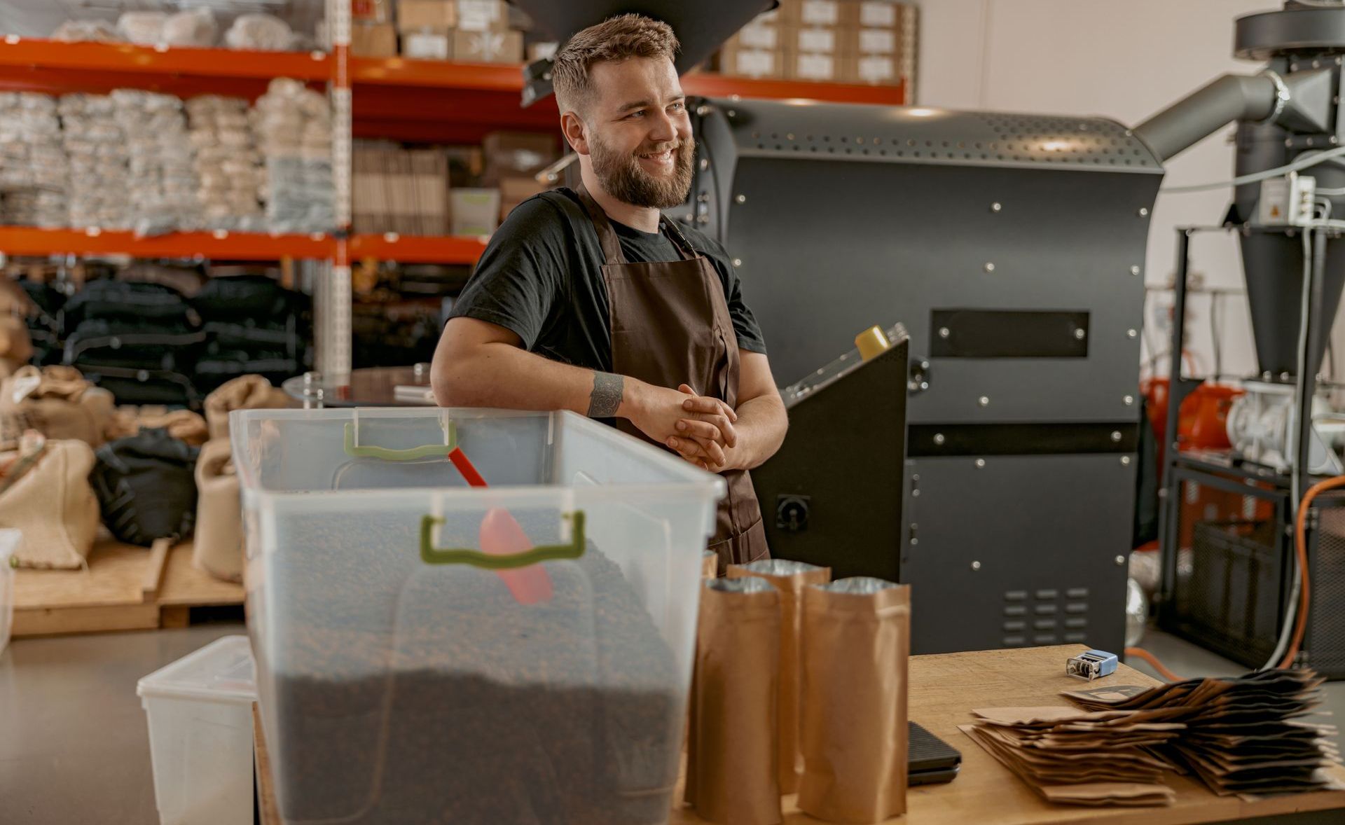 A man is standing in front of a coffee roaster in a warehouse.