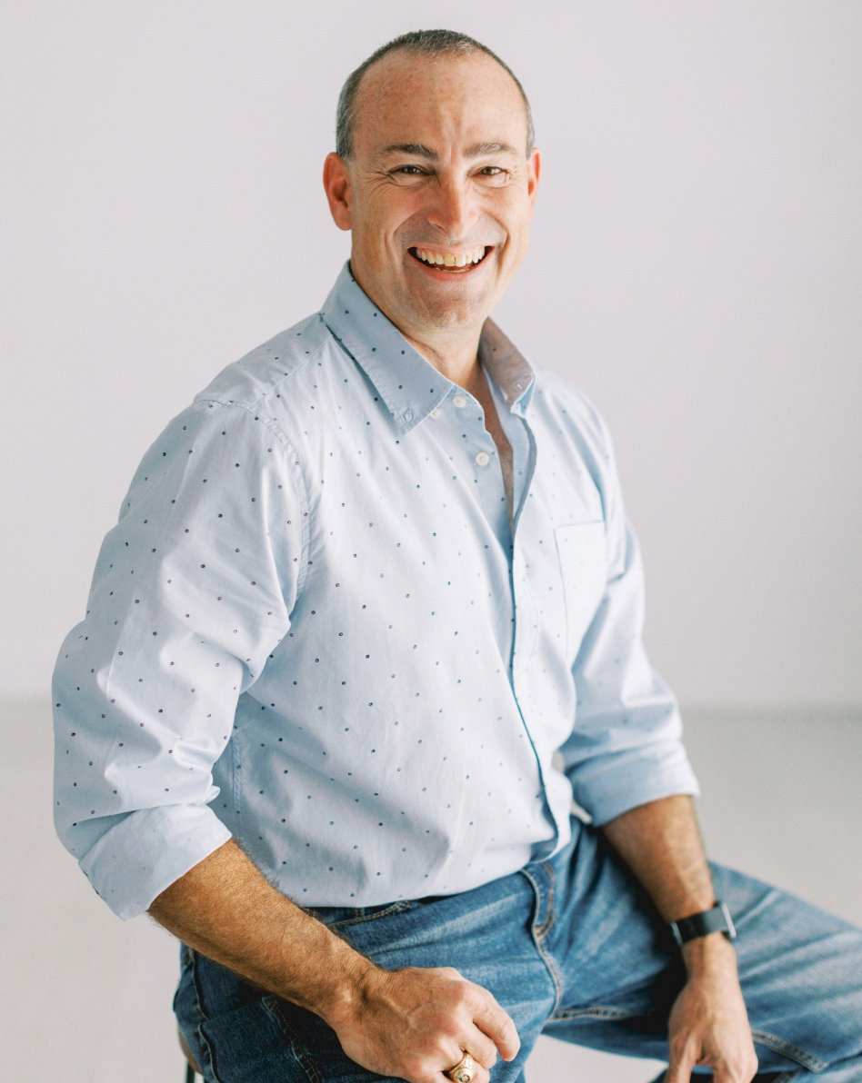 A man in a blue shirt and jeans is sitting on a stool and smiling.