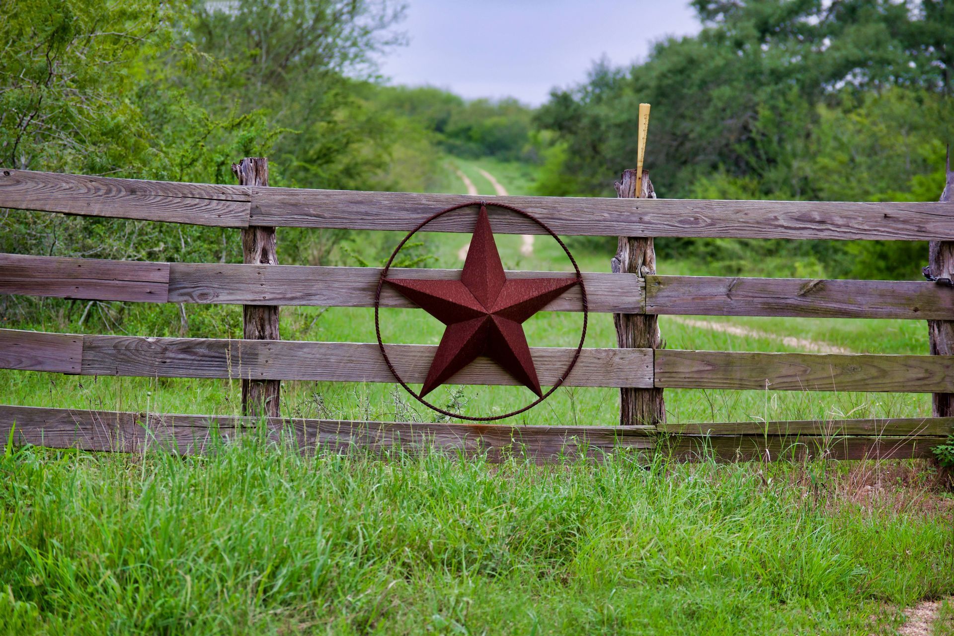 Wooden fence with rusty star decoration, leading to a dirt path in a green field.