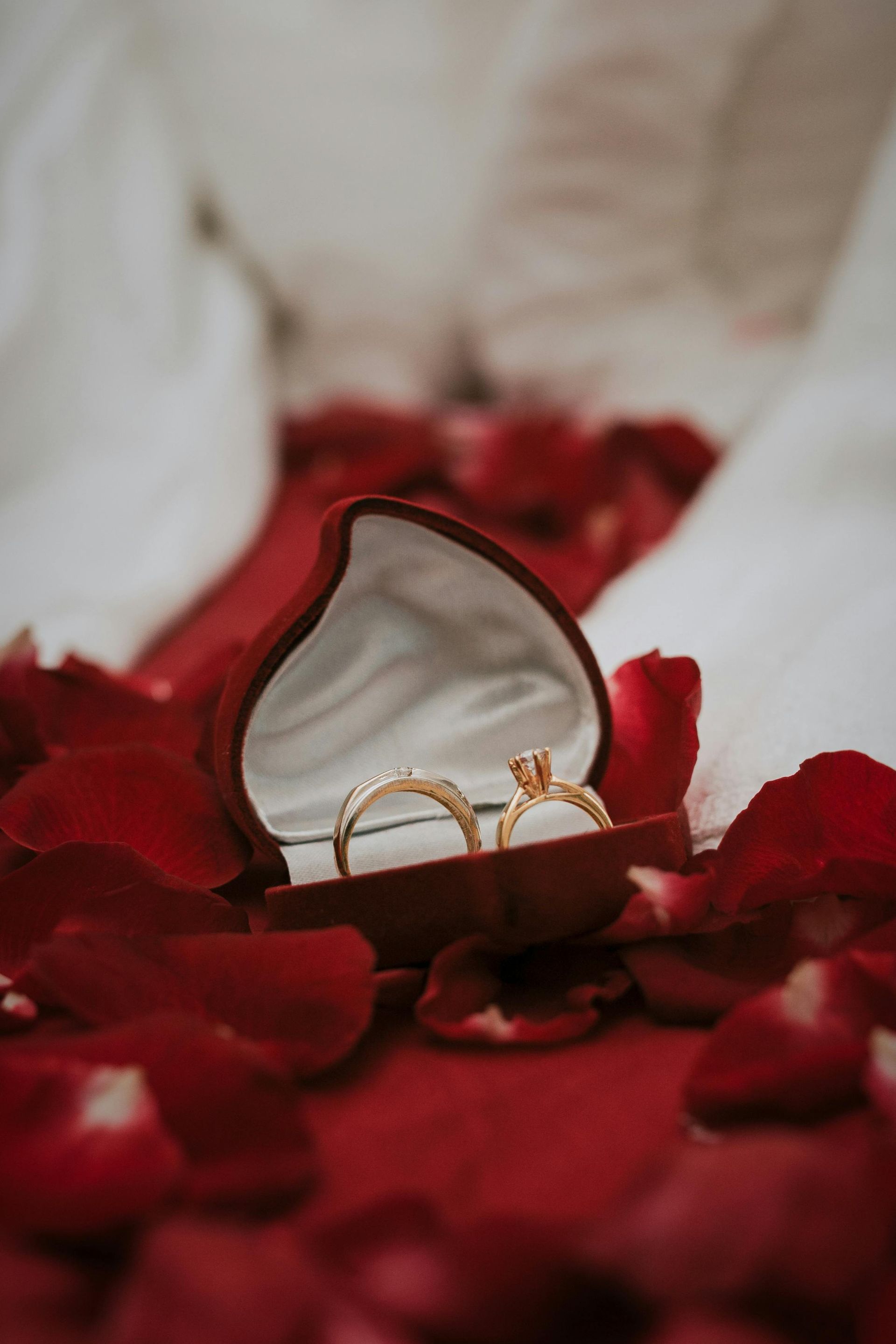 A pair of wedding rings in a heart shaped box surrounded by rose petals.