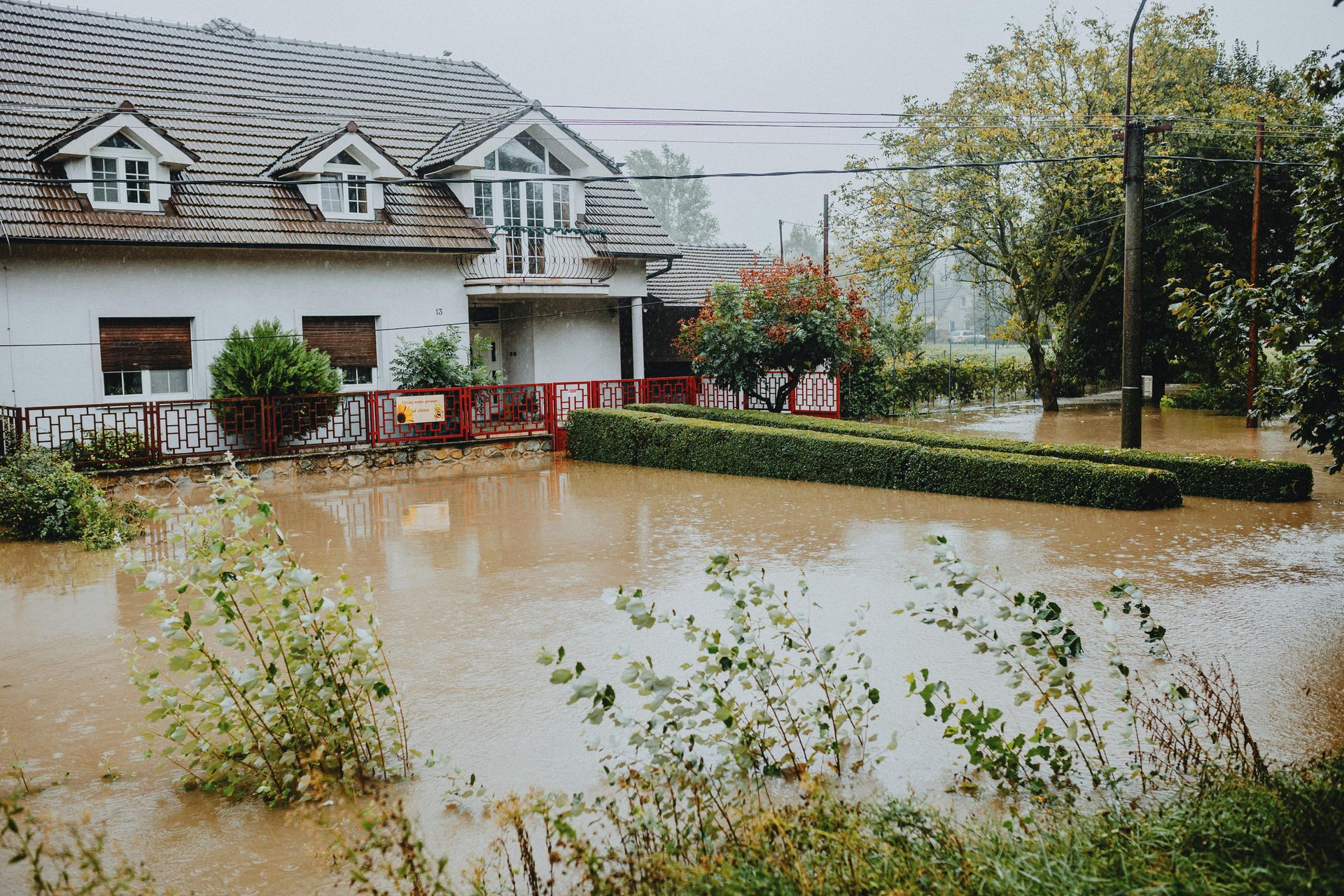 Flooded house. Water surrounds a two-story home with a white exterior and a tiled roof, under a cloudy sky.