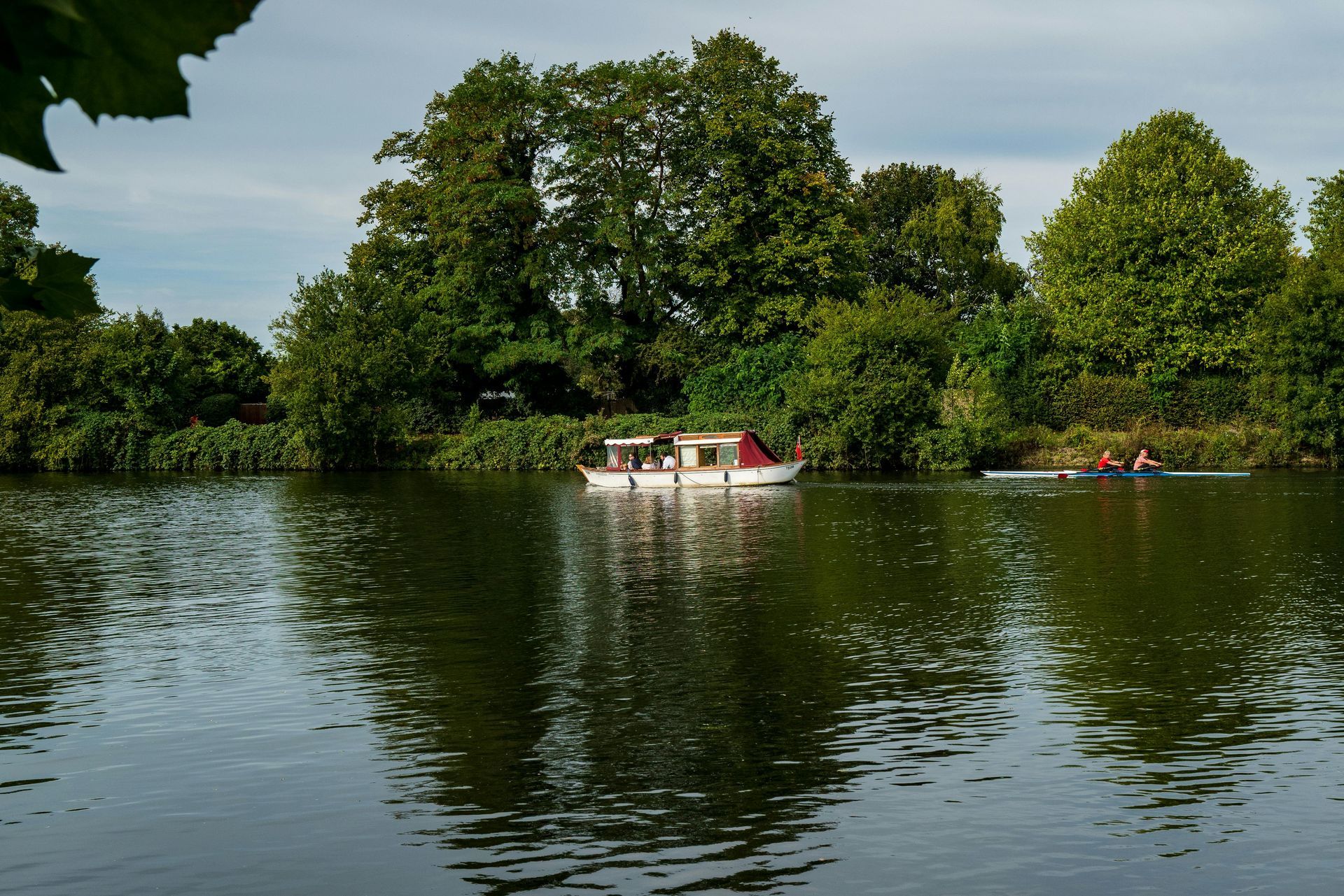 River scene with a motorboat and rowers on the water, surrounded by green trees under a cloudy sky.