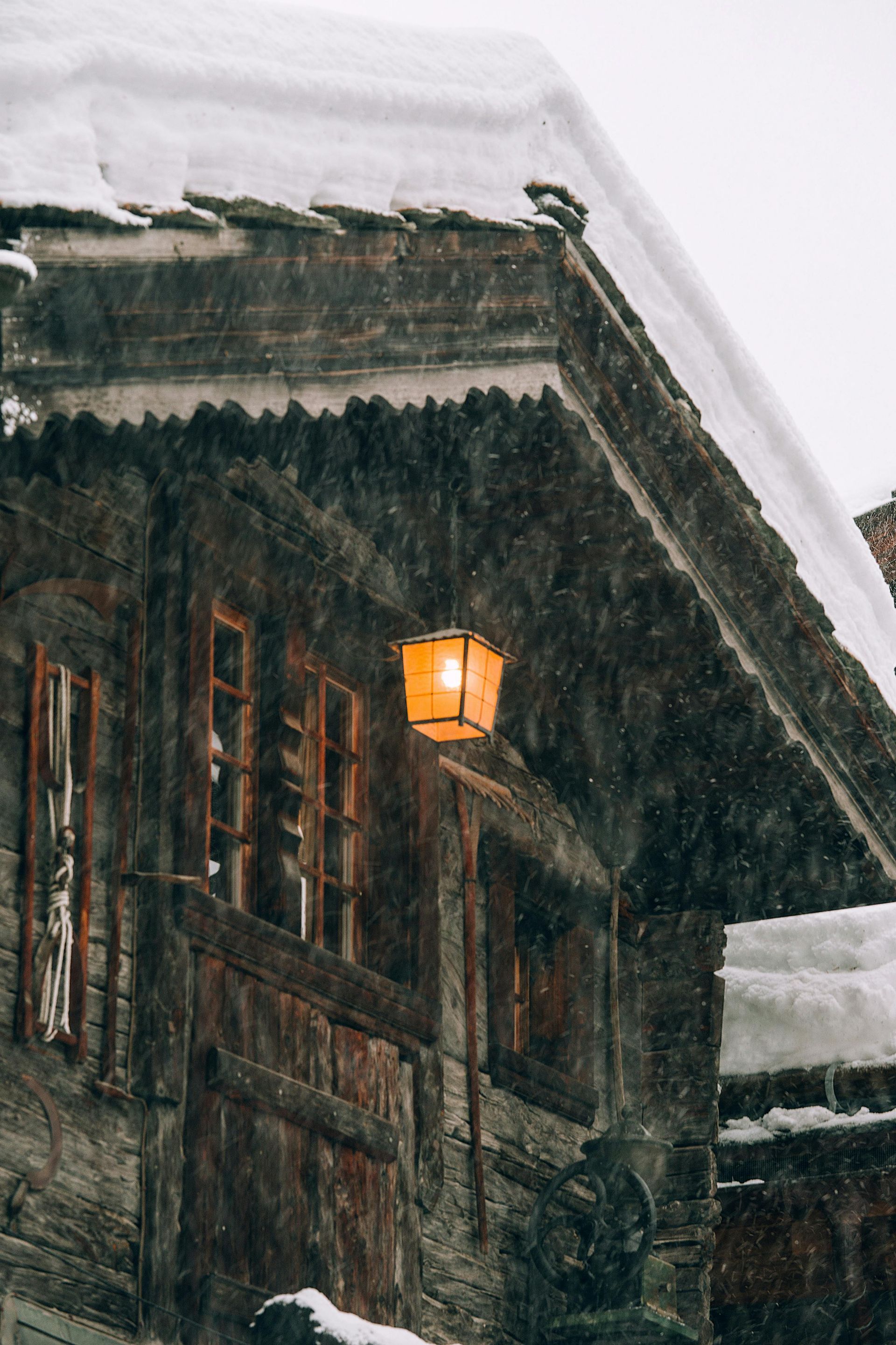 Snowy scene of an old wooden building with an illuminated orange lantern. Snow on roof, windows visible.