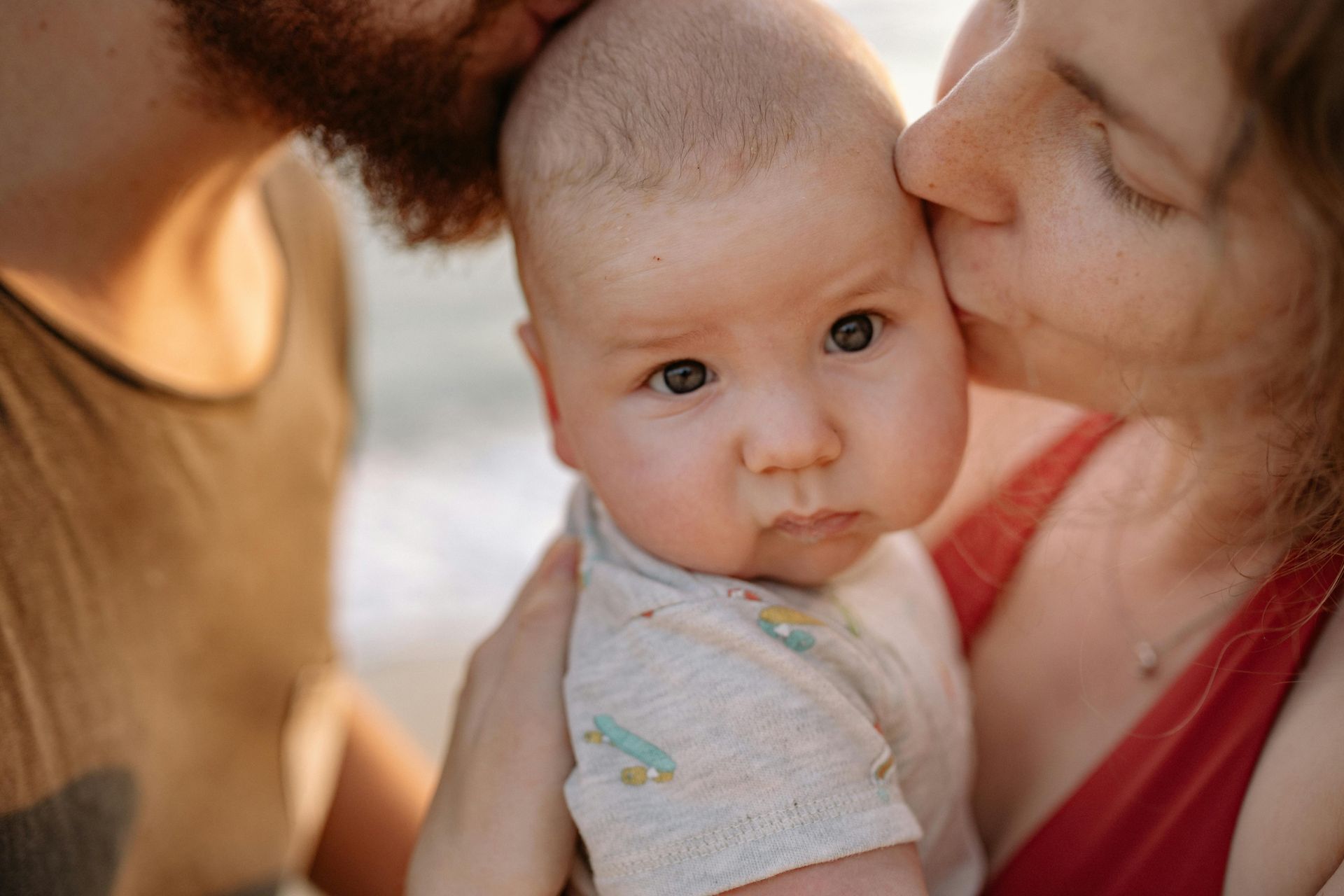 Baby held by parents, kissed on the cheek, outdoors. Baby has a neutral expression, parents close. Warm light.