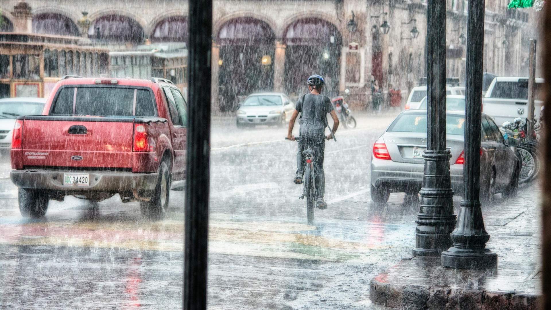 Cyclist rides bike in heavy rain on city street with cars; red pickup truck in front.