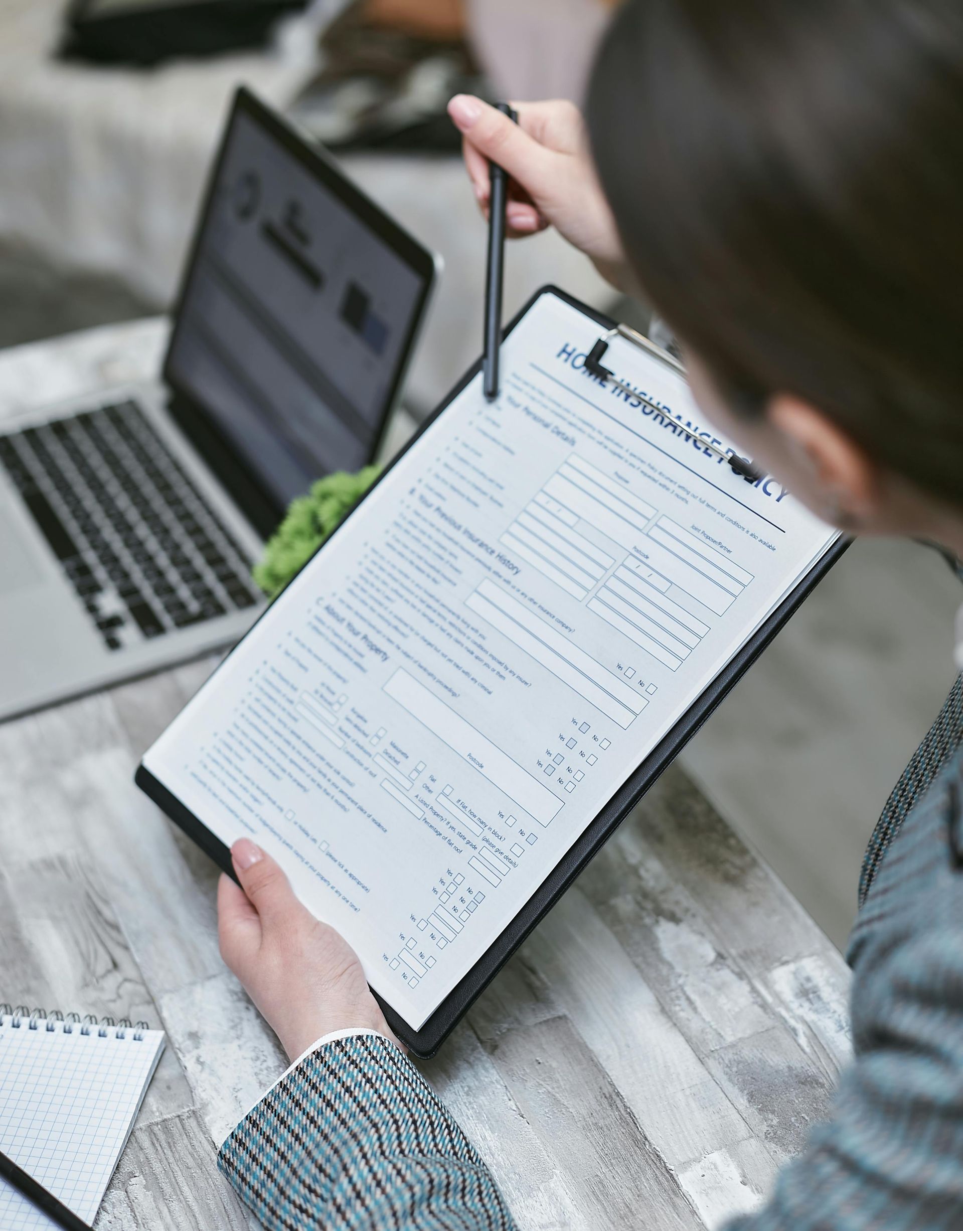 Person reviewing a clipboard beside a laptop, holding a pen over paperwork.