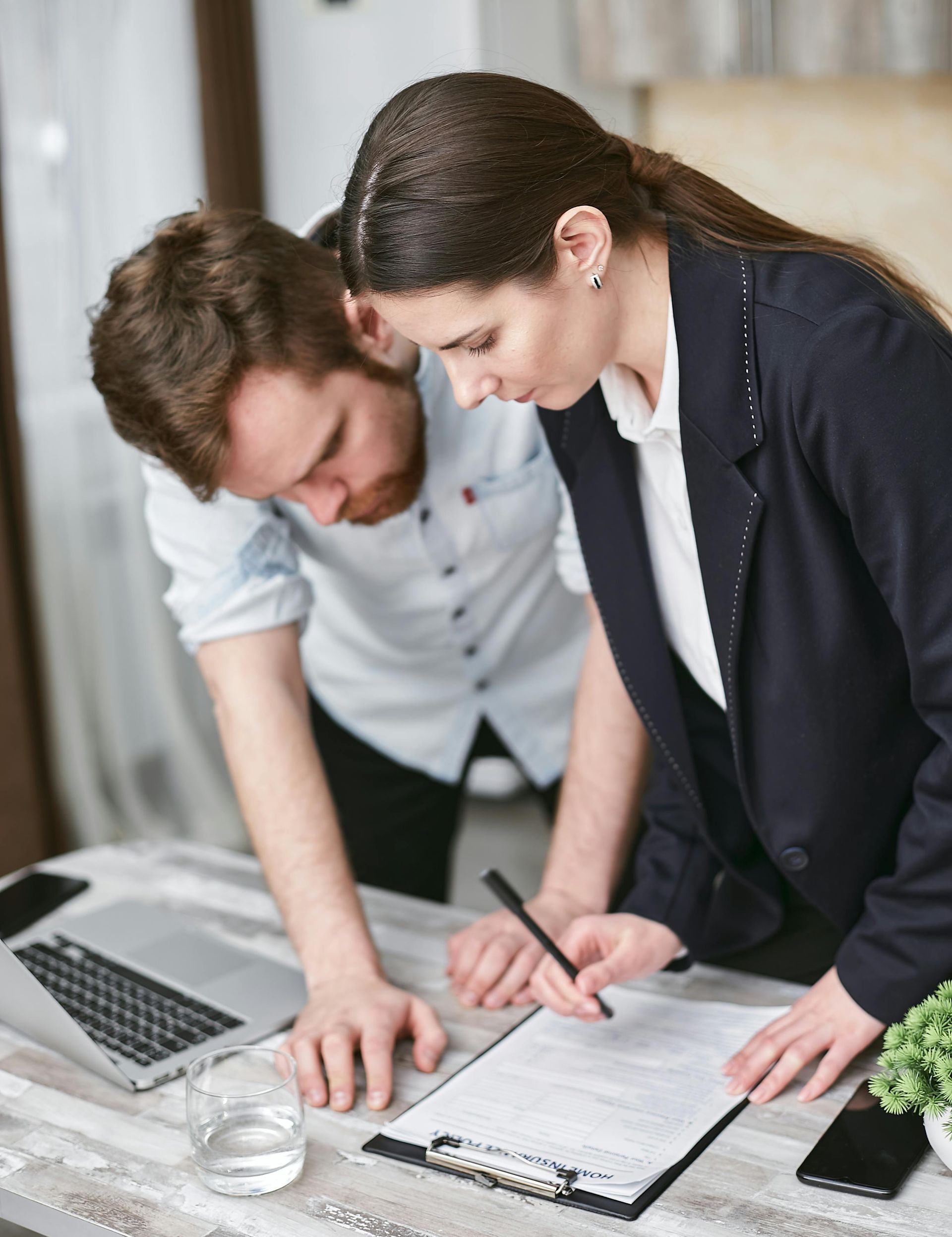 Woman in blazer pointing at document, man leans in. They are at a desk with a laptop and glass.