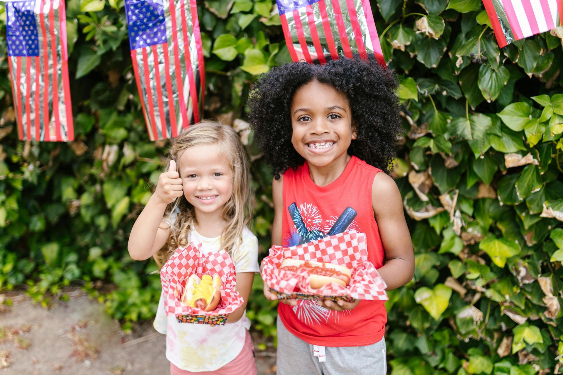 Two children smiling, holding hotdogs, with American flags in the background.