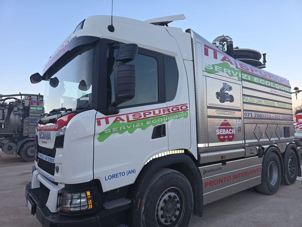 Un grosso camion aspiratore argentato con segnaletica rossa e verde è parcheggiato all'aperto su un piazzale ghiaioso.