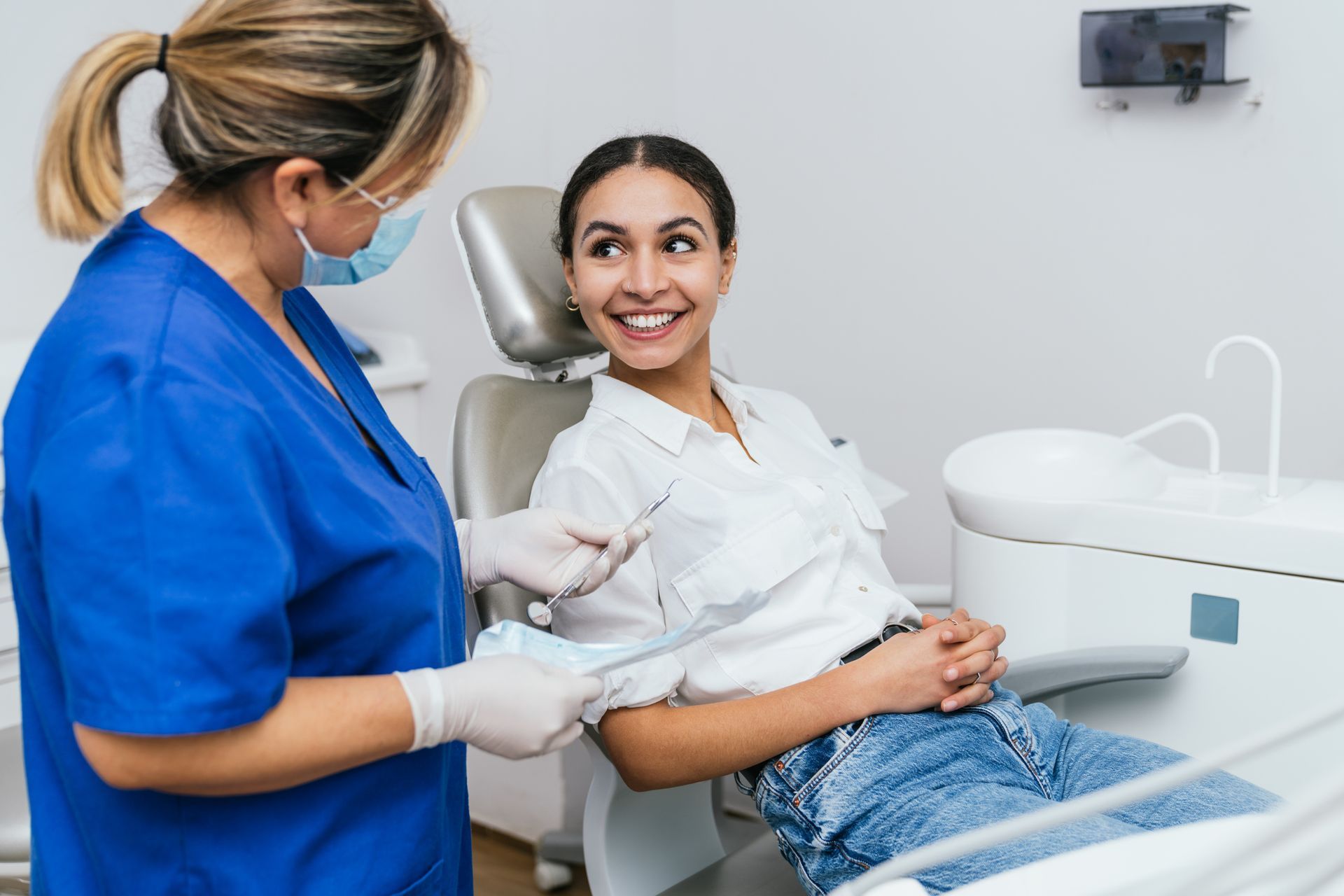 A female dentist with blue clothing is attending to a smiling young woman in her dental office.