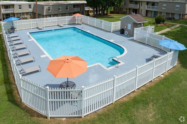 Swimming pool surrounded by white fence, lounge chairs, and umbrellas, in a grassy area near apartment buildings.