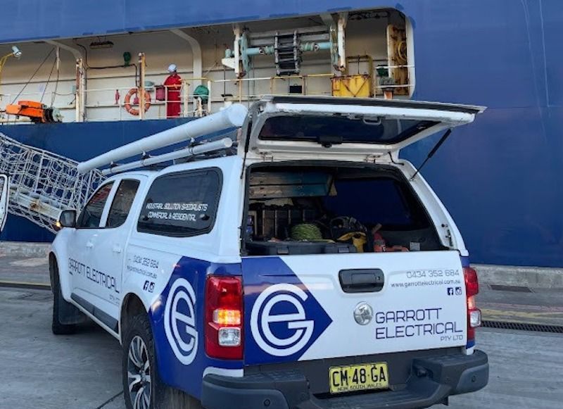 A white and blue Garrott Electrical utility vehicle parked with its tailgate open near a large ship.
