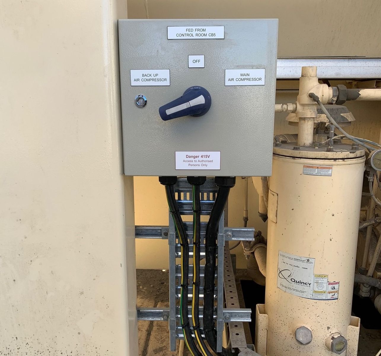 Gray electrical control box with a blue dial switch mounted next to a vertical industrial pipe and tank.