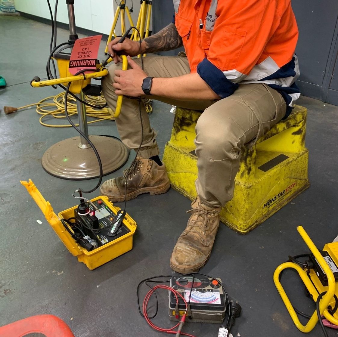 A worker wearing high-visibility orange clothing sits on a yellow step stool, testing electrical equipment on a floor.