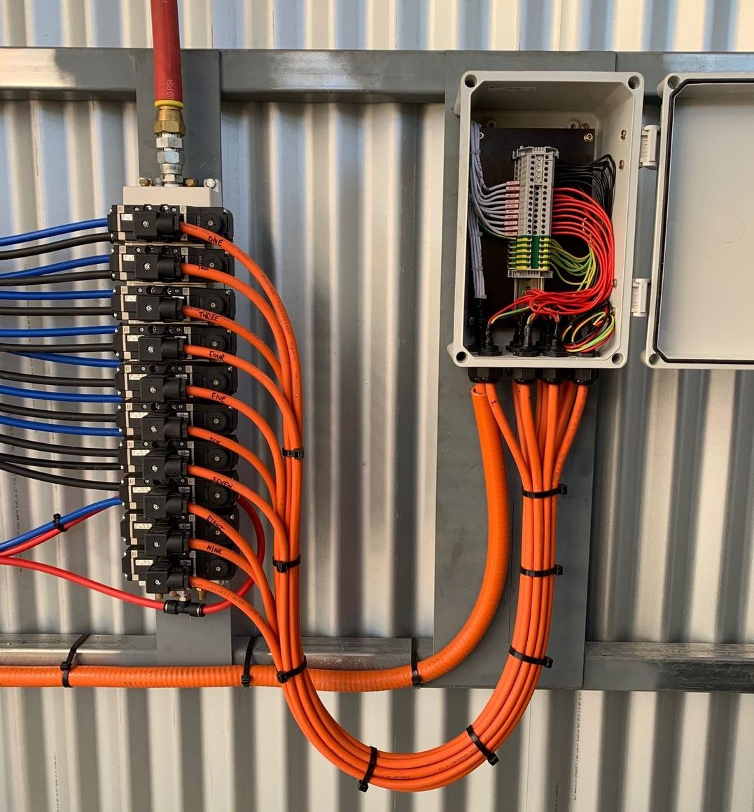A pneumatic manifold with orange tubing and a wired electrical junction box mounted on a corrugated metal wall.