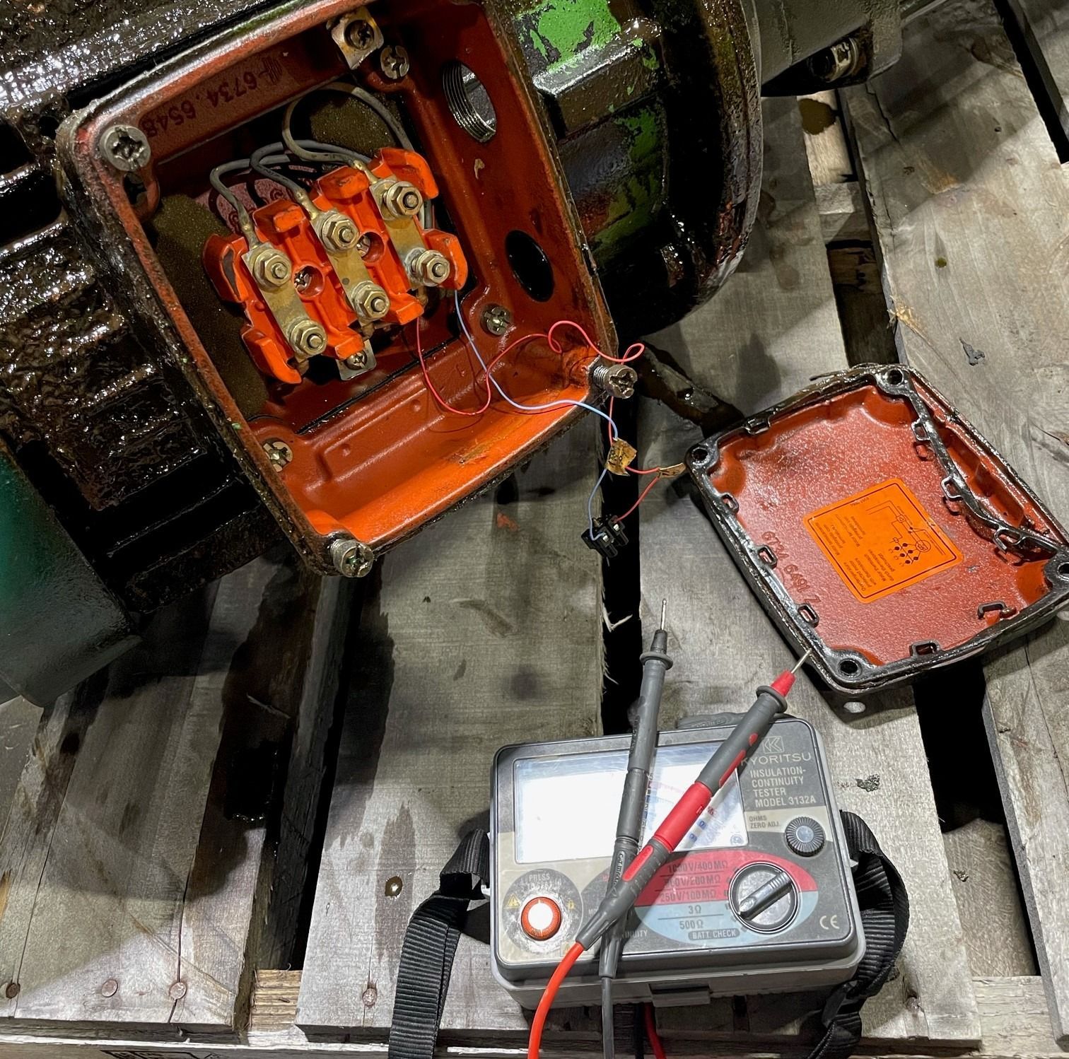 A multimeter with probes resting on a wooden surface next to an open electric motor terminal box with orange components.