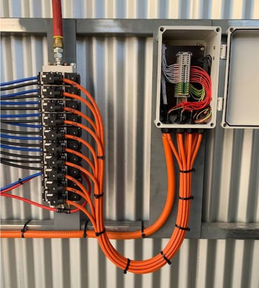 A solenoid valve manifold with orange cables connects to an open junction box on a corrugated metal wall.