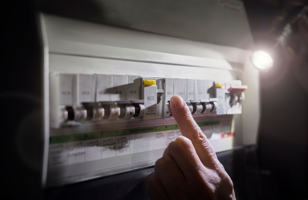 A person's hand reaches toward an electrical consumer unit to flip a tripped circuit breaker switch in dim light.