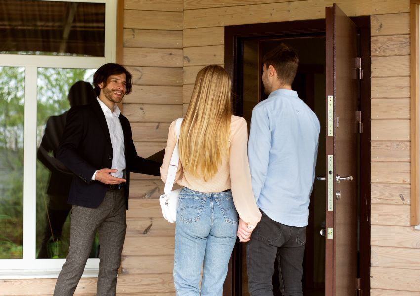 A smiling property manager in a black suit jacket and white dress shirt ushers two prospective tenants into a wooden paneled property for a tour.