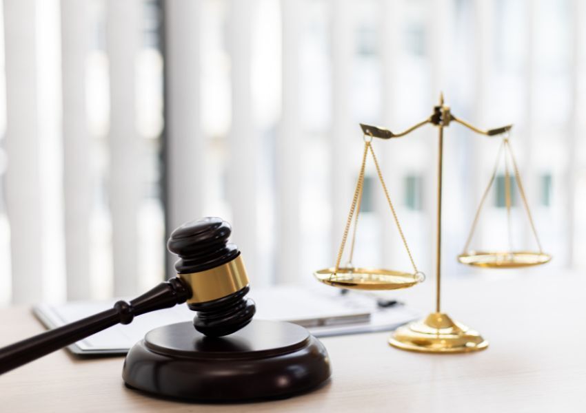 A dark brown and gold gavel is pictured on a white desk, next to a gold balancing scale.