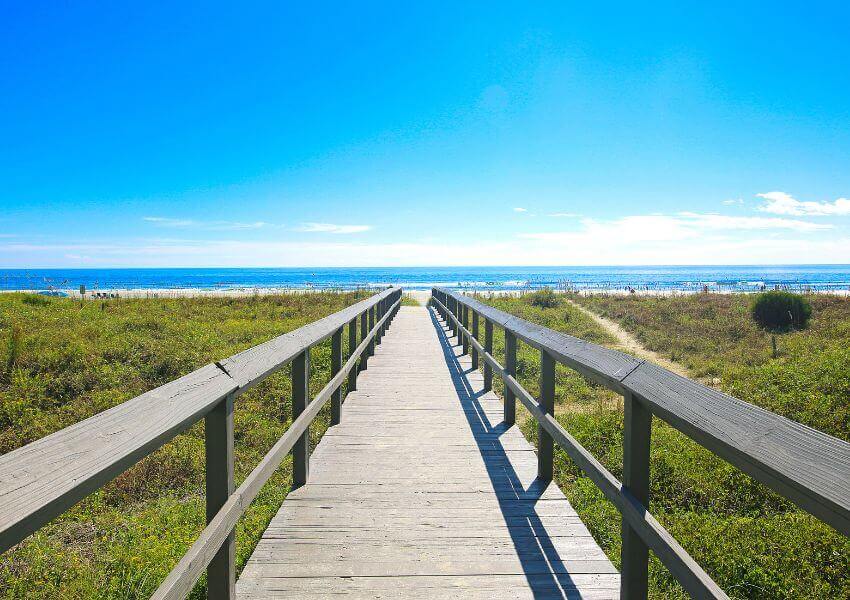 A scenic view of wooden walkway leads to a beach with grass that meets the sand and water.