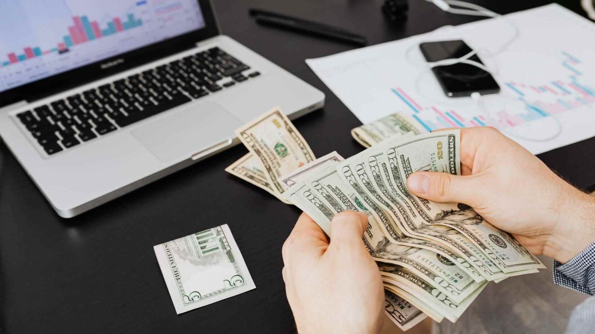 A close-up on a property investor’s hands counting American money at a desk, with figures on a laptop in the background.