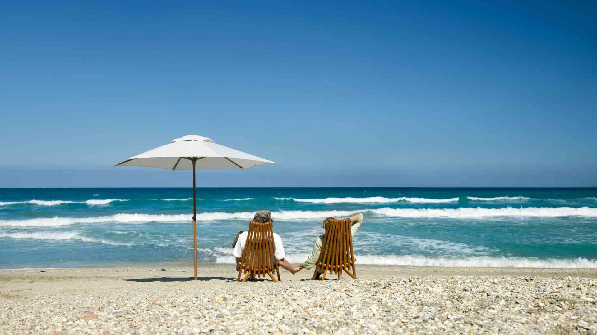 A retired couple sitting beside an umbrella on wooden chairs, shot on a sandy beach with calm waves and a cloudless sky.