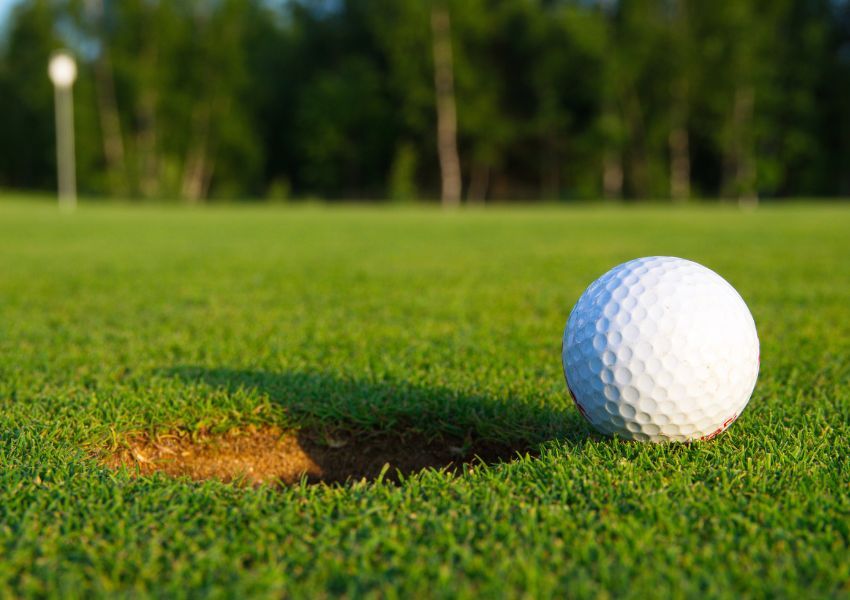 A close up of a white golf ball sitting beside the dirt hole on perfectly trimmed, short grass.